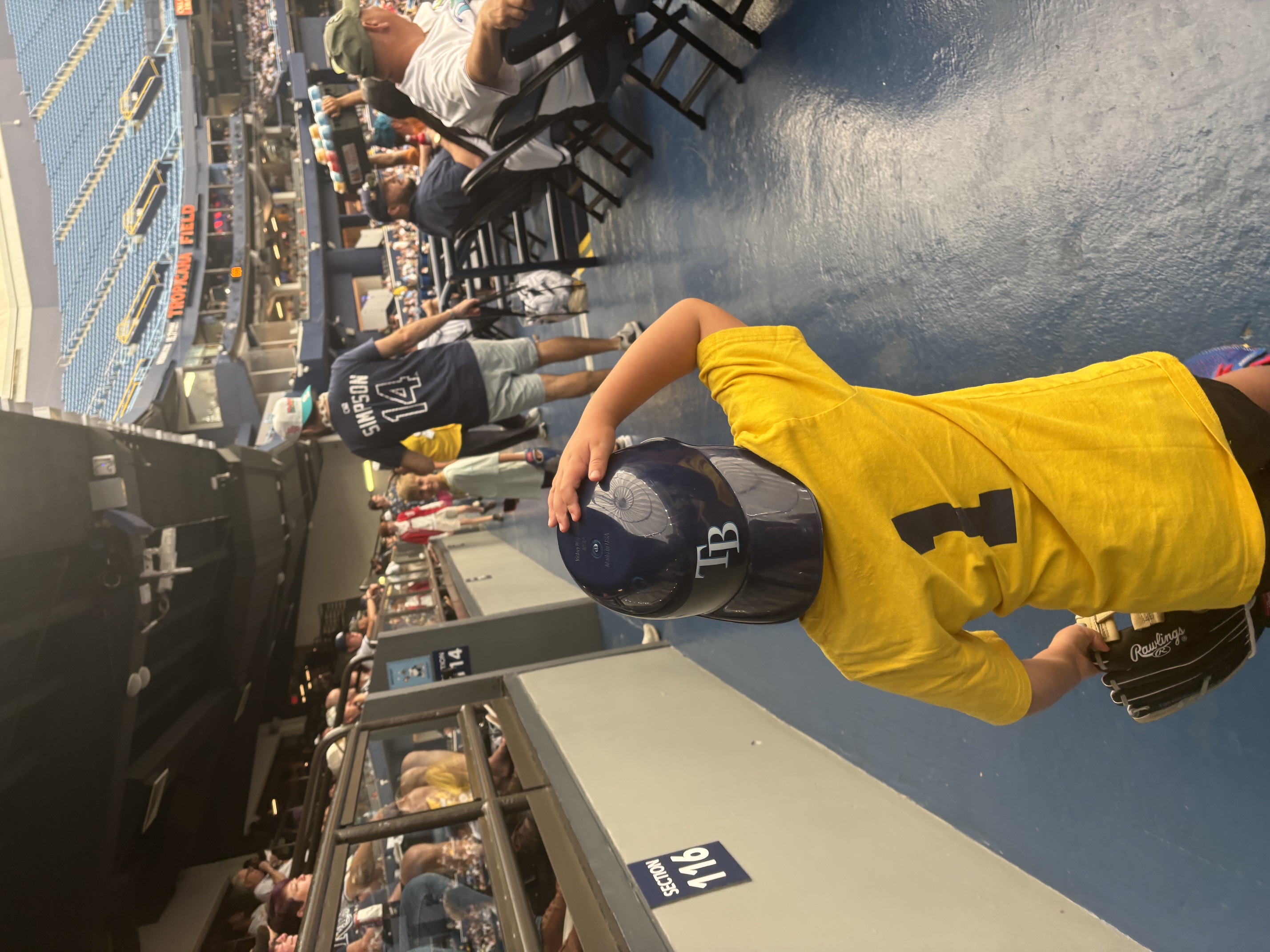 A boy in a yellow number 1 shirt holding a Rays batting helmet at the railing